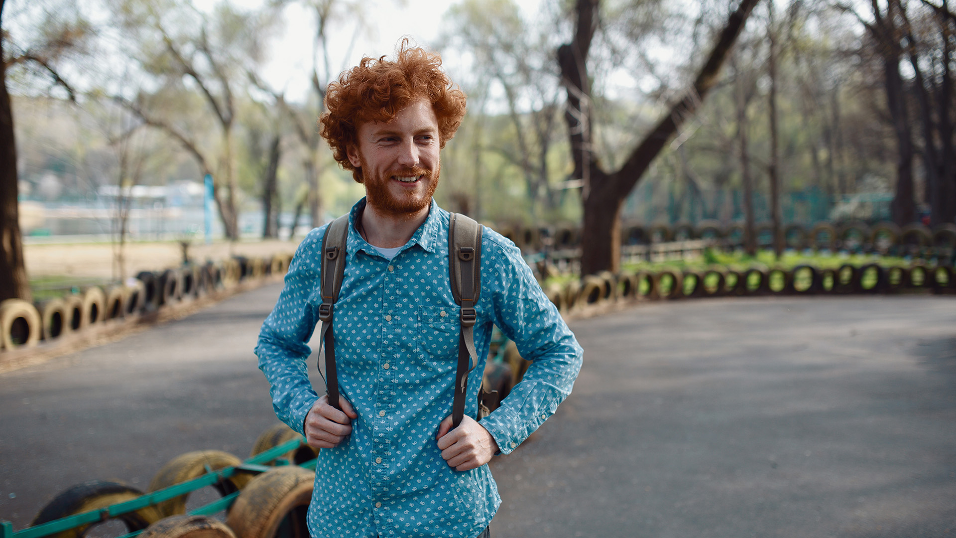 Man med rött hår , blå skjorta och grön ryggsäck står utomhus och ler och tittar åt sidan.