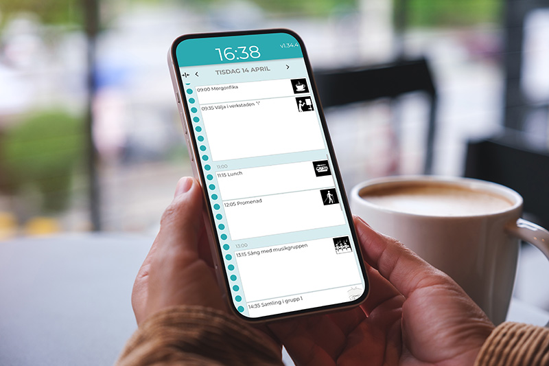 Två händer håller en telefon med en dagsvy av Boet för daglig verksamhet. Bredvid står en kopp kaffe.