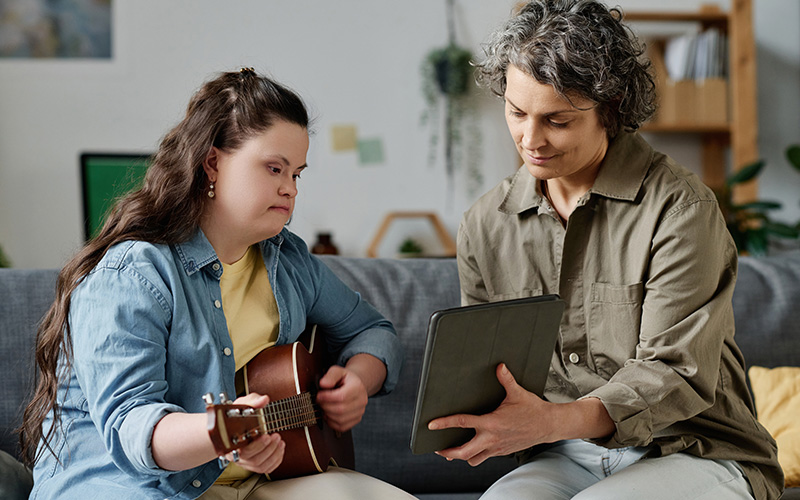 Två kvinnor sitter i en soffa på ett LSS-boende, den ena spelar gitarr och den andra håller upp en ipad framför.