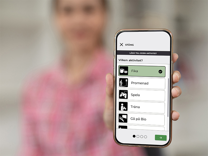 En hand håller upp en mobiltelefon som visar Boets funktion "Lägg till egen aktivitet", med valbara alternativ som Fika, Promenad, Spela, Träna och Gå på bio.