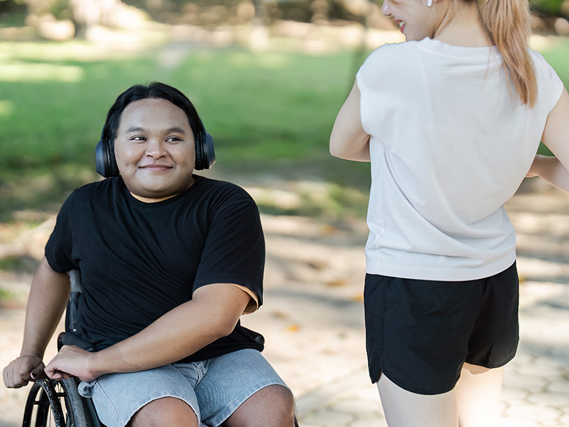Man i rullstol med svart t-shirt, short och hörlurar tränar tillsammans med en kvinna som står i vit t-shirt och svarta shorts.
