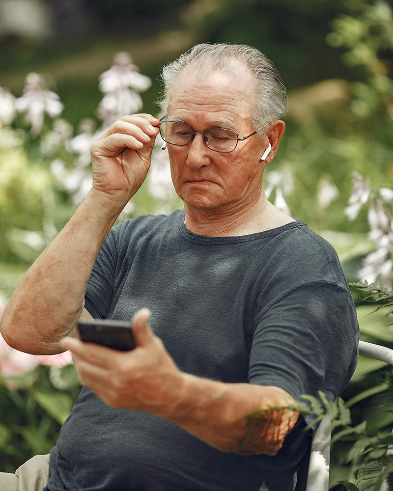 Äldre man med glasögon och airpods tittar ner i sin smartphone med grönska bakom sig.